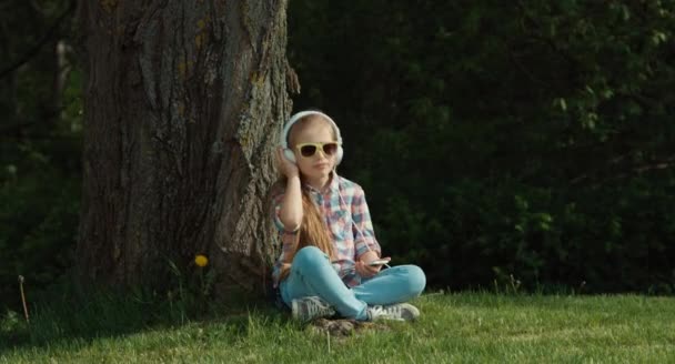 Κορίτσι ακούγοντας μουσική. Το παιδί κάθεται κάτω από ένα δέντρο. Της τα χέρια το τηλέφωνο — Αρχείο Βίντεο
