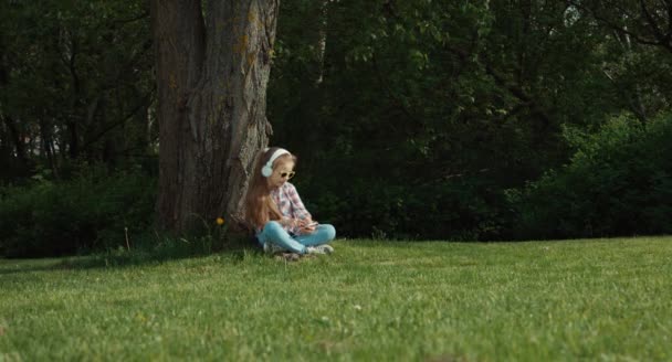 Dívka, poslechu hudby. Dítě sedící pod stromem. V její ruce telefon. Dítě je v režimu online — Stock video