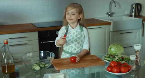 Criança cortando os tomates e colocá-los na tigela de salada. Menina chef na cozinha olhando para a câmera e sorrindo — Vídeo de Stock