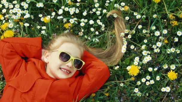 Lachendes Mädchen, das im Gras liegt und in die Kamera blickt. Mädchen in rotem Mantel und Sonnenbrille. Kind träumt und tut nichts. Zoomen — Stockvideo