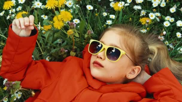 Lachendes Mädchen, das eine Blume riecht und im Gras liegt und in die Kamera blickt. Mädchen in rotem Mantel und Sonnenbrille. Mädchen mit Blumen. Zoomen — Stockvideo