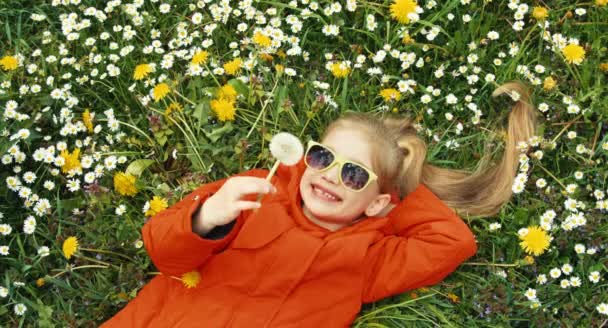 Closeup portrait of a child. Girl holding a dandelion and laughing at the camera. The child lies on the grass among flowers — Stock Video