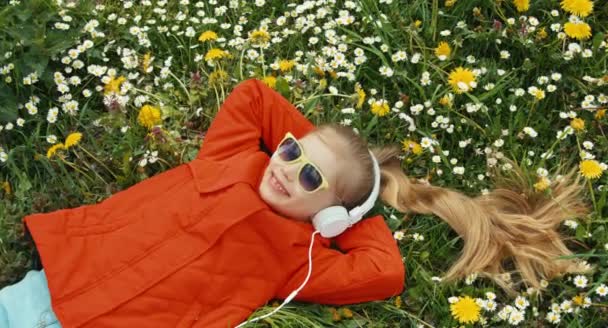 Menina retrato close-up ouvindo música. A criança deitada na grama entre as flores. Panning — Vídeo de Stock
