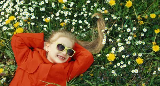 Gülen kız çim yalan ve kameraya bakıyor. Kız kırmızı ceket ve güneş gözlüğü. Rüya çocuk ve hiçbir şey yapmaz — Stok video