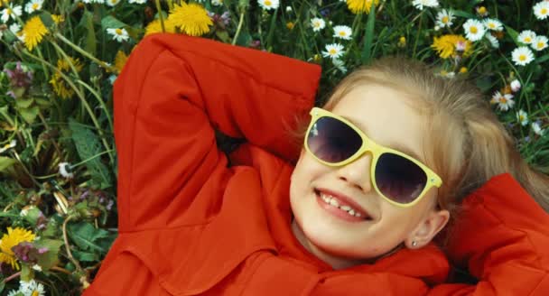 Muchacha retrato de primer plano acostada en la hierba y mirando a la cámara. Chica con un abrigo rojo y gafas de sol — Vídeos de Stock