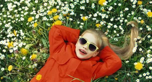 Une fille qui chante une chanson et rit. Gros plan portrait enfant couché dans l'herbe et regardant la caméra dans des lunettes de soleil. Panoramique — Video