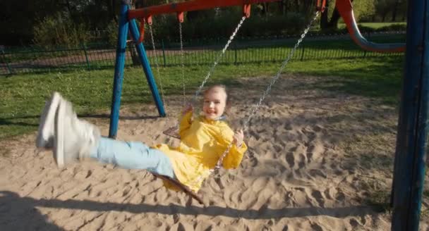 Menina montando em um balanço no parque e olhando para a câmera — Vídeo de Stock