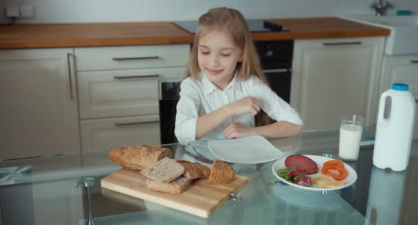 Portrét dívky preschooler v kuchyni. Poblíž dítě je bochník chleba, salát, sýr, klobása a sklenici mléka. Dívka bude připravit sendvič. Ona konzumní mléko — Stock video