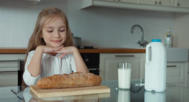 Retrato de una niña preescolar en la cocina. Cerca del niño hay una barra de pan y un vaso de leche. De acuerdo, pulgares arriba. — Vídeos de Stock