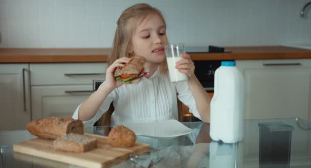 Retrato de una niña preescolar en la cocina. Chica sosteniendo un sándwich y bebiendo leche. Sonriendo a la cámara — Vídeo de stock