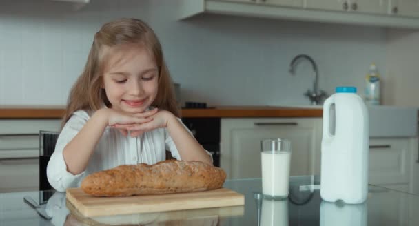 Retrato de una niña preescolar en la cocina. Cerca del niño hay una barra de pan y un vaso de leche. De acuerdo. Pulgar hacia arriba — Vídeos de Stock