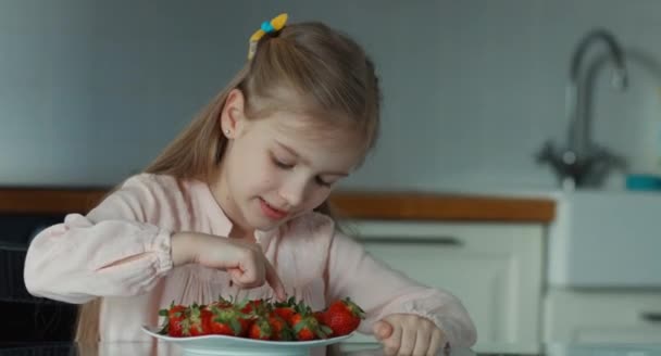 Primer plano retrato niño riendo y mirando a la cámara. Pulgares arriba. Junto a ella hay un gran plato de fresas. Chica contando fresas — Vídeos de Stock