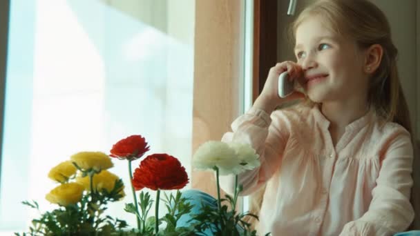 Little girl talking on cell phone sitting on the windowsill. She laughing and looking at camera — Stock Video