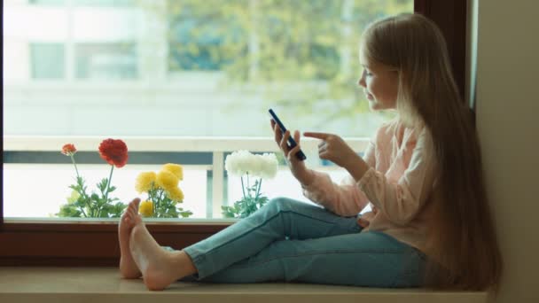 Niño usando el teléfono móvil y sonriendo. Mirando la cámara. Muestra bien, pulgares arriba. Niño sentado en la ventana alféizar — Vídeos de Stock