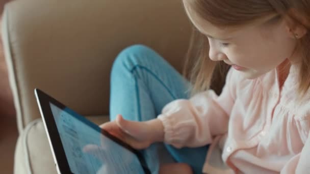 Primo piano Bionda bambina è online con tablet pc e seduta sul divano. Bambino ridendo e guardando la fotocamera — Video Stock