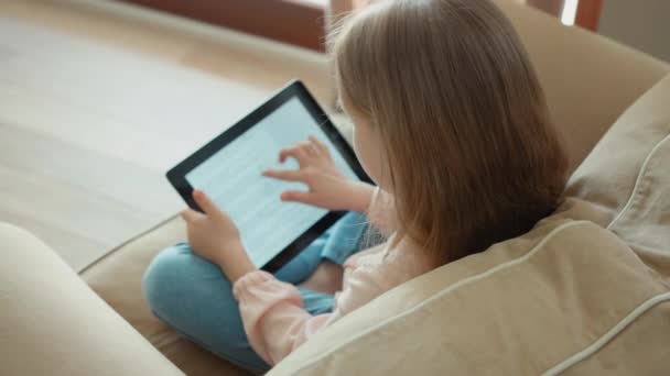 Blond dziewczyna siedzi na kanapie i przy użyciu komputera typu tablet. Dziecko jest w trybie online — Wideo stockowe