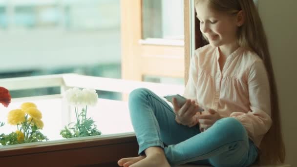 Niño usando el teléfono móvil y sonriendo. Mirando la cámara. Muestra bien, pulgares arriba. Chica sentada en la ventana alféizar — Vídeo de stock