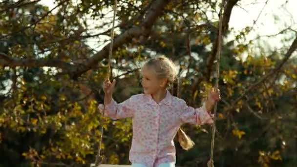 Joyeux enfant sur une balançoire sous un arbre. Fille regardant caméra et sourire — Video