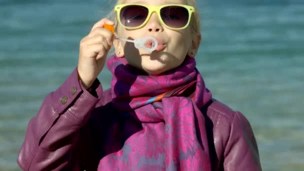 Closeup portret van een meisje in zonnebril bubbels op het strand van zee waait. Kind kijken camera — Stockvideo