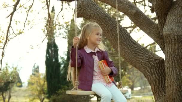 Miling girl hugging the books and looking at camera. Child sitting on a swing — Stock Video