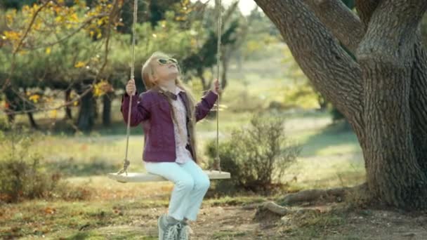 Niña preescolar en gafas de sol mirando al cielo con placer en un columpio — Vídeos de Stock