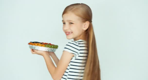 Stående flicka håller kakan med frukt nära ansiktet på den vita bakgrunden — Stockvideo