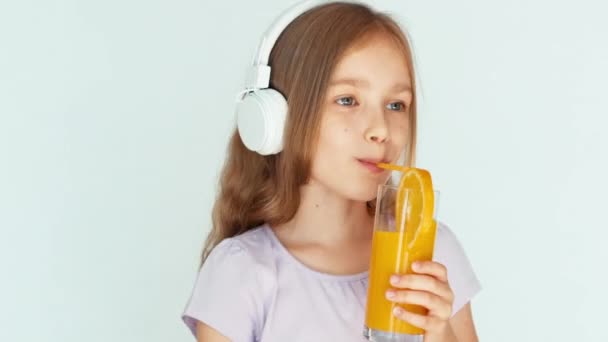 Meisje drinken sinaasappelsap en dansen en muziek koptelefoon luisteren en glimlachend in de camera. Meisje met mooie blonde haren op een witte achtergrond. Closeup — Stockvideo