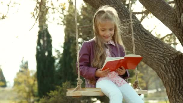 Школярка читає червону книгу і дивиться на камеру. Дитина сидить на гойдалках — стокове відео