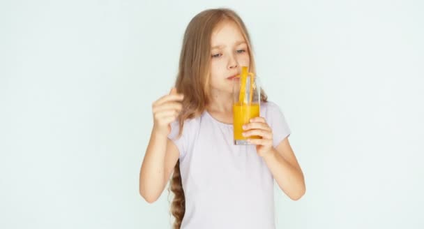 Chica bebiendo jugo de naranja y bailando y sonriendo a la cámara. Chica con hermoso cabello rubio sobre un fondo blanco. Pulgar hacia arriba. Ok. — Vídeos de Stock