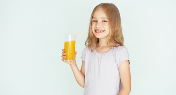 Chica bebiendo jugo de naranja y sonriendo a la cámara. Chica con hermoso cabello rubio sobre un fondo blanco. Niño asiente. Pulgar hacia arriba. Ok. — Vídeos de Stock