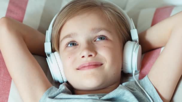 Vértes portré lány gyermek zenehallgatás fejhallgató és az ágyon pihent és mosolyogva kamera. Kicsinyítés/nagyítás — Stock videók