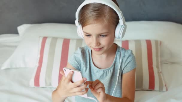 子供のベッドの上に座ってスマート フォン携帯電話とダンスからヘッドフォンで音楽を聴きます。親指を。わかりました。ズーム — ストック動画