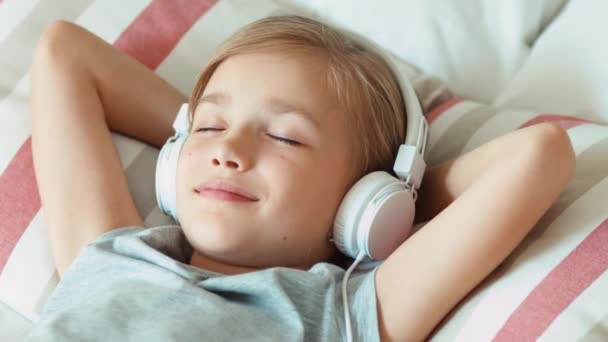 Primer plano niña retrato escuchando música en los auriculares con los ojos cerrados y acostado en la cama y descansando y sonriendo a la cámara. Ampliación — Vídeos de Stock