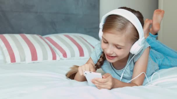 Дитина лежить на ліжку. Дівчина слухає музику в навушниках і використовує мобільний телефон. великий палець вгору Добре. Збільшення — стокове відео