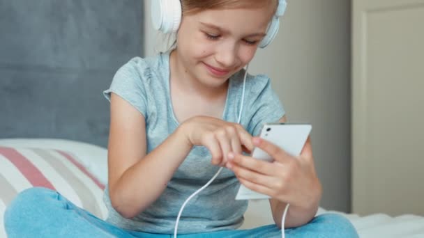 Niña sentada descansando en la cama y escuchando música en auriculares desde un teléfono celular y bailando y riéndose de la cámara — Vídeos de Stock