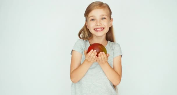 Девушка держит и показывает манго и смеется над камерой. Ребенок с фруктами на белом фоне — стоковое видео