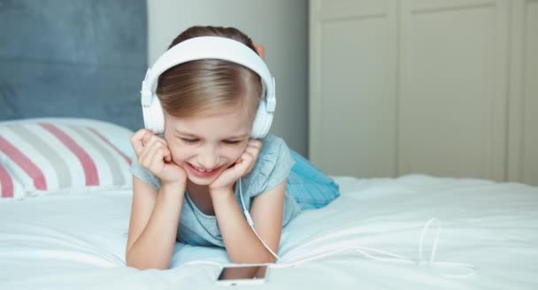 Крупним планом портретна дівчина слухає музику в навушниках і співає пісню. Дитина лежить на ліжку і сміється з камери — стокове відео