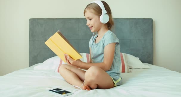 Дівчина слухає музику в навушниках і читає обіймаючу книгу. Дитина сидить на ліжку і посміхається — стокове відео