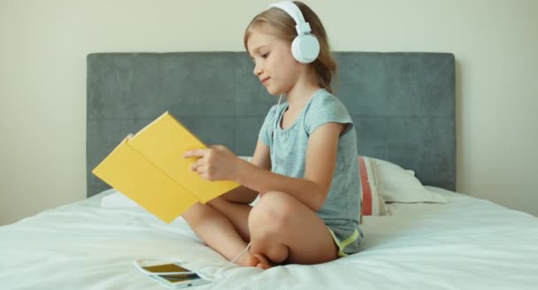 Meisje luisteren muziek in hoofdtelefoons en leesboek. Kind zitten rusten op het bed en glimlachend op camera — Stockvideo