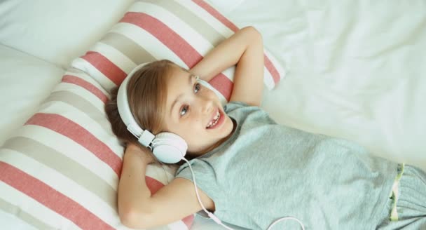 Chica escuchando música en auriculares. Niño acostado descansando en la cama y riendo de la cámara — Vídeo de stock
