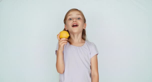 Mädchen beißt in eine Zitrone und hat ein geschwungenes Gesicht. Kind lacht in die Kamera — Stockvideo