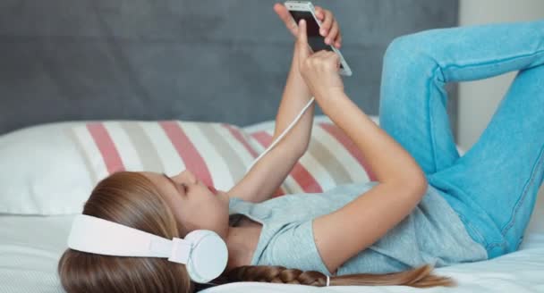 Девушка лежит на кровати, слушает музыку в наушниках и использует смартфон — стоковое видео