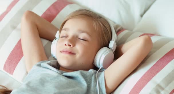 Primer plano retrato niña escuchando música en los auriculares con los ojos cerrados y acostado en la cama — Vídeos de Stock