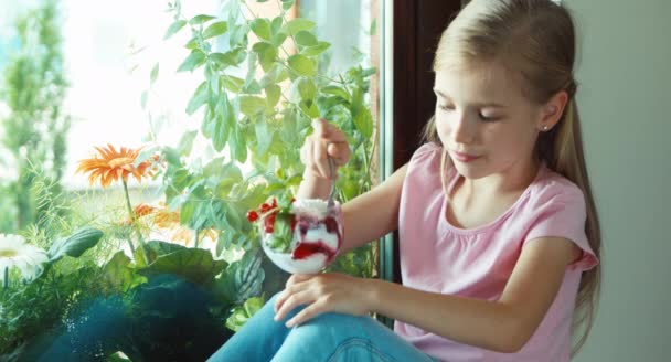 Girl eating ice cream against window and smiling at camera. The child nods at the camera — Stockvideo