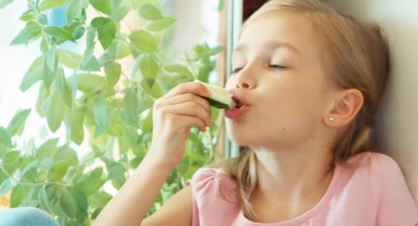 Closeup portret meisje komkommer eten tegen raam — Stockvideo