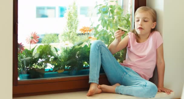 Meisje komkommer eten en zitten op de vensterbank — Stockvideo