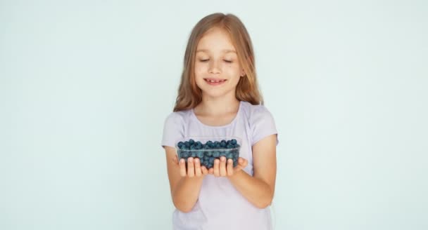 Girl sniffing blueberry and showing at camera on the white background — Stockvideo