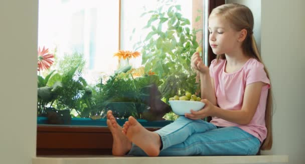 Mädchen, das auf einer Fensterbank sitzt, Stachelbeeren isst und in die Kamera blickt. Daumen hoch. ok — Stockvideo