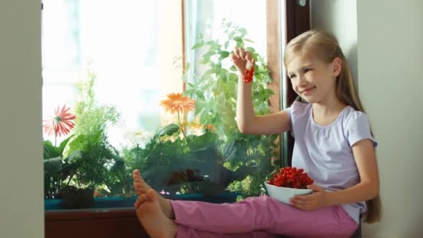 Girl sniffing very tasty sweet red currants and smiling at camera. Child sitting on the windowsill. Zooming — Stock video