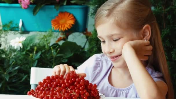 Meisje met een takje van rode aalbessen. Meisje heeft een grote plaat van rode bessen. Kind zitten rustend op de tafel in de tuin. Zoomen — Stockvideo